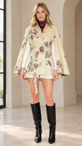 Front view of woman wearing beige floral botanical belted cape coat with tall black boots