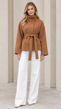 Front view of model wearing camel stand collar belted short coat with white trousers